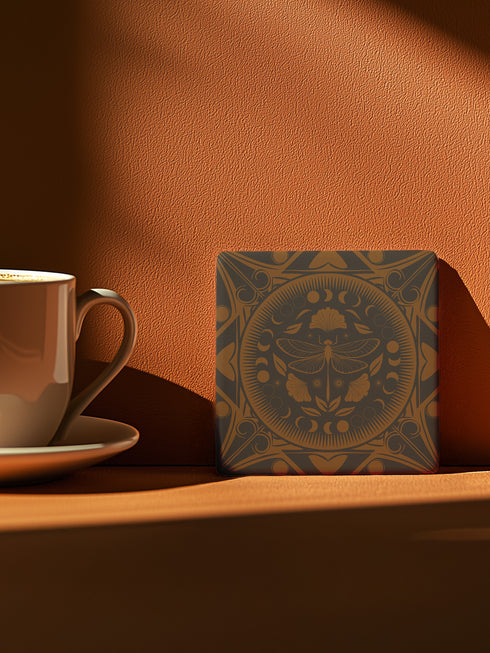 Square ceramic coaster featuring a detailed copper dragonfly design centered in a medallion, surrounded by moon phases and Art Nouveau patterns on a charcoal black background, with cork backing.